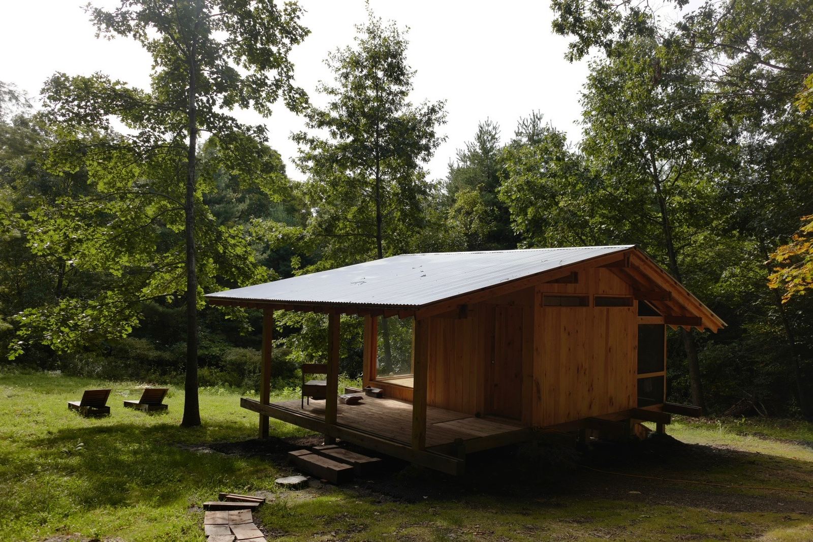 The timber cottage under a corrugated metal roof, with a deep covered porch, in a sunny clearing ringed by trees